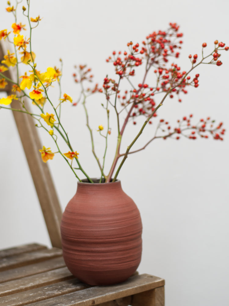 Handgemaakte rode vaas met bloemen erin, op een houten stoel met witte achtergrond