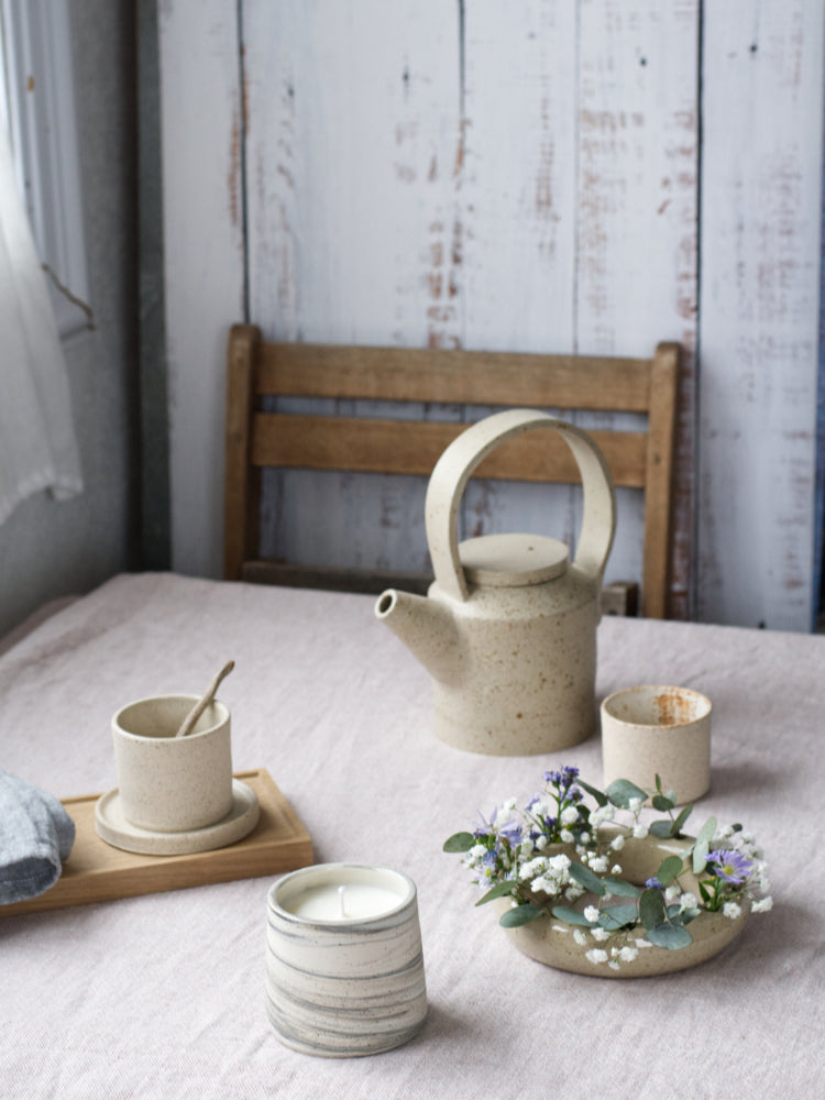 Ronde bloemenvaas op een gedekte tafel met handgemaakte theepot, kopjes en kaars.