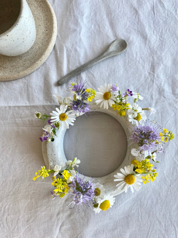 Ronde handgemaakte bloemenvaas met paarse, gele en witte bloemen op een tafelkleed met een handgemaakt lepeltje van keramiek ernaast