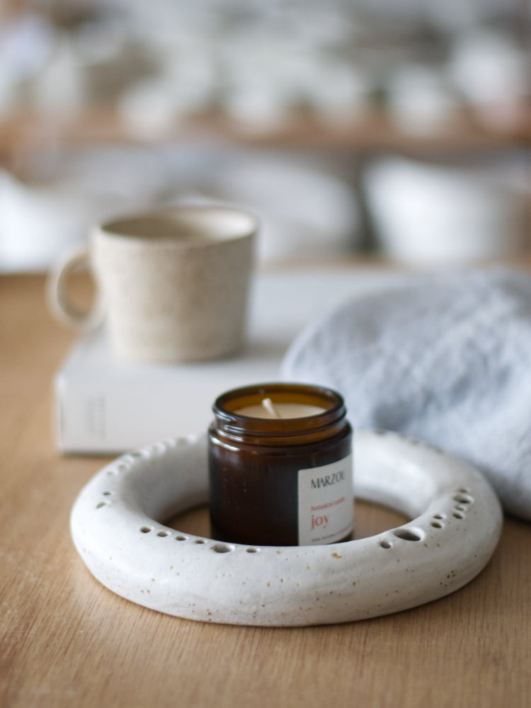 Ronde handgemaakte vaas met Joy geurkaars op houten tafel, met vaag een boek, mok en blauwe theedoek op de achtergrond