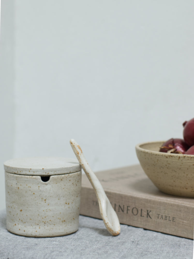 Handgemaakt suikerpotje van keramiek met een lepel ertegenaan naast een boek met een kommetje erop.