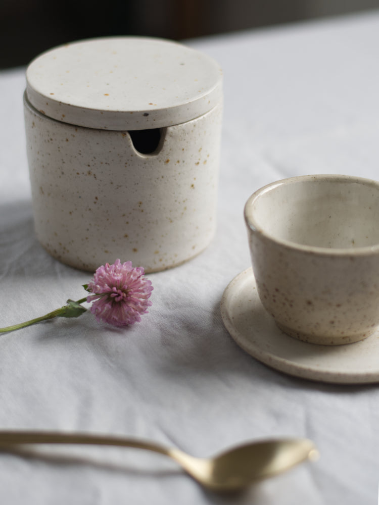 Suikerpotje handgemaakt, op een tafelkleed met een kopje en schotel, bloemetje ernaast, japandi stijl.