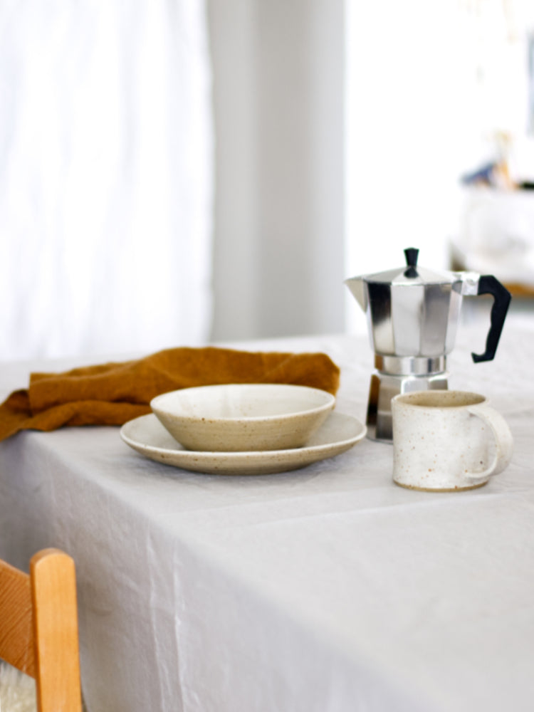 Een gedekte tafel met tafelkleed, een stoel en handgemaakt keramiek servies met een percolator ernaast
