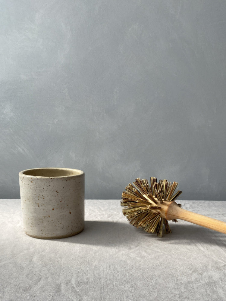 Handgemaakte toiletborstelhouder van gespikkeld keramiek met ernaast een houten borstel, op een lichtgrijze achtergrond