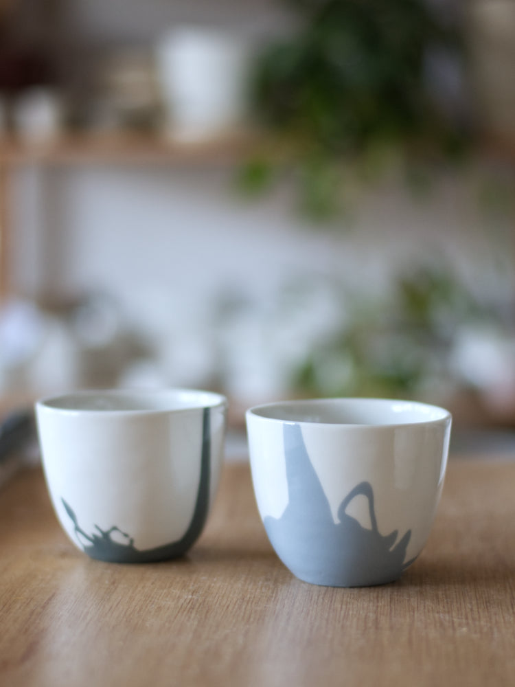 Twee handgemaakte Waterlijn kopjes van wit porselein met een zachte gekleurde waterlijn, staand op een houten tafel met hangplant vaag op de achtergrond.