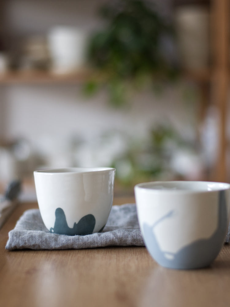 Twee handgemaakte Waterlijn kopjes van wit porselein met een zachte gekleurde waterlijn, staand op een houten tafel met een linnen doek op de achtergrond.