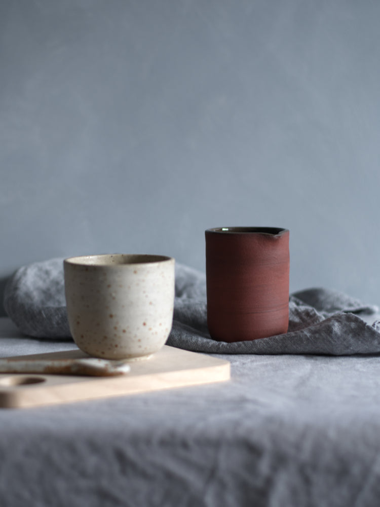 Rode schenkkan, naast een handgemaakt kopje van gespikkelde klei, met een grijze achtergrond, heel stijlvol, op een grijsblauw tafelkleed met een blauwe theedoek erbij.