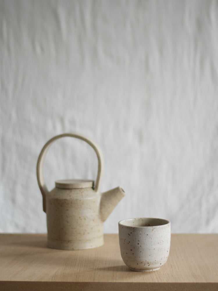 Whitewashed kopje, op een houten ondergrond, voor een handgemaakte theepot. De theepot is vaag in de achtergrond.