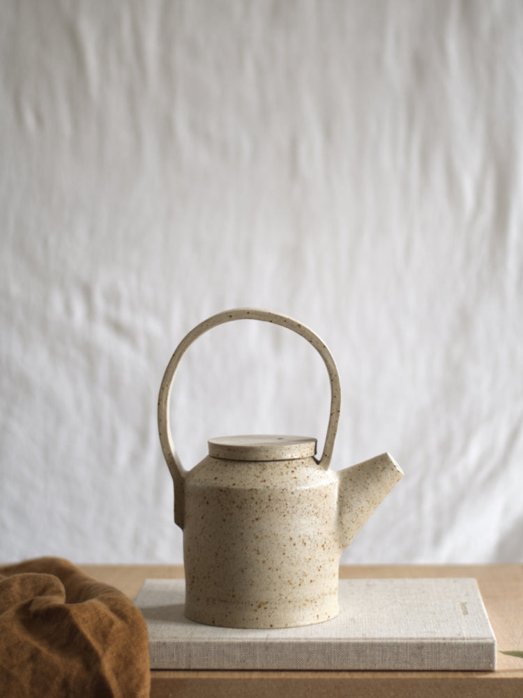 Handgemaakte theepot van gespikkeld keramiek, op een boek met een theedoek ernaast. Slow living sfeer 