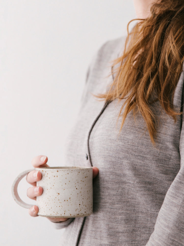 Handgemaakte mok van gespikkelde klei die wordt vastgehouden door een vrouw met roodbruin haar. Haar gezicht is niet te zien