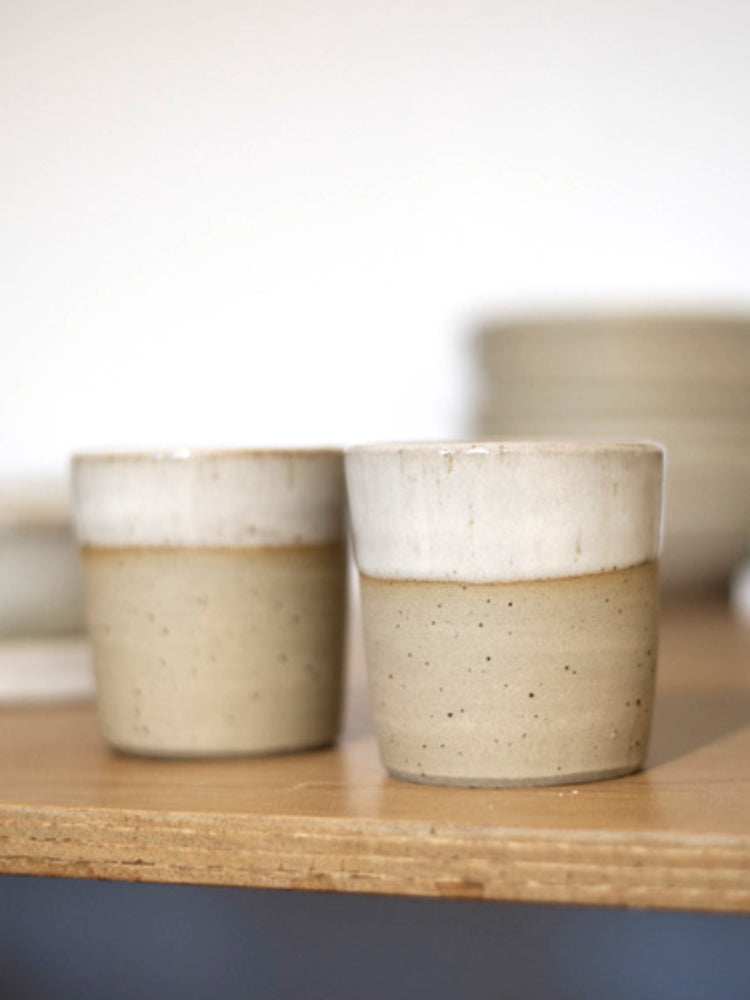 Twee handgemaakte bekers van gespikkeld steengoed met glanzend wit glazuur, waarbij de spikkels van de klei zichtbaar blijven en de bovenrand geglazuurd is
