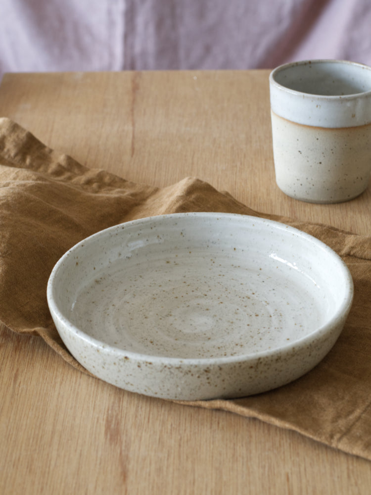 handgemaakte keramieken kom op bruine  theedoek met een handgemaakte beker erachter op een houten tafel