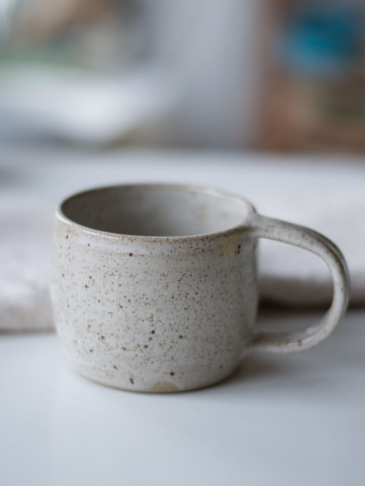 Handgemaakte mok wit gespikkeld glazuur naast een theedoek op een witte tafel, closeup