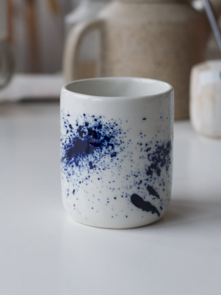 Handgemaakte wit porseleinen kopje met blauwe spetters op een witte tafel, close up gefotografeerd