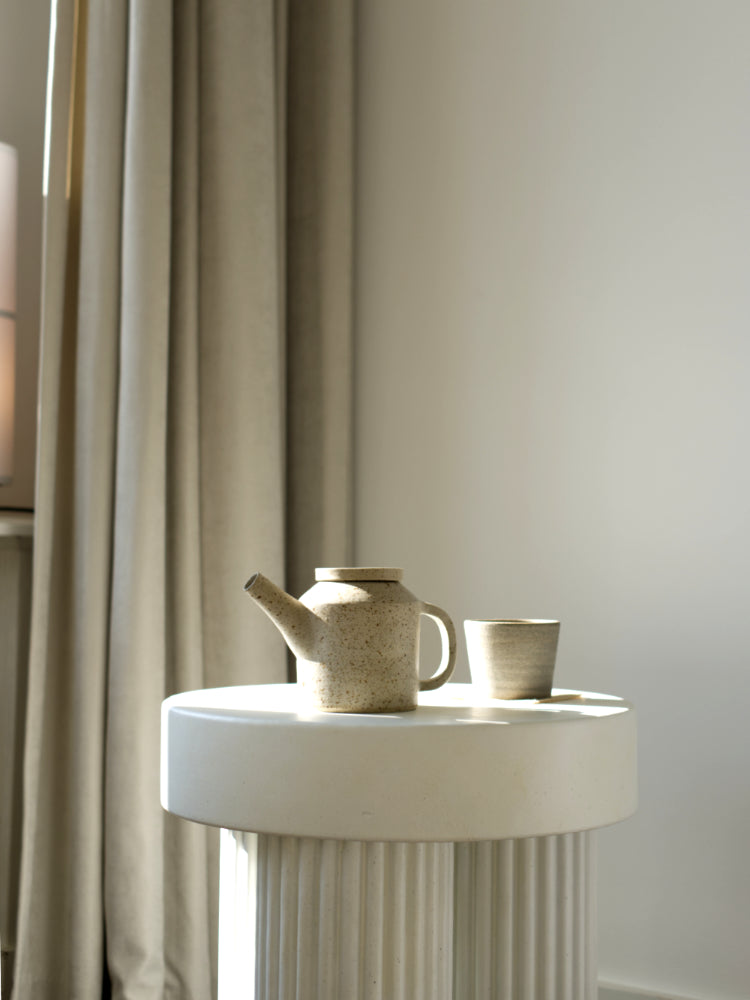 Rond laag tafeltje met daarop een handgemaakte theepot en kopje van gespikkelde klei in zonlicht, op de achtergrond lange gordijnen 