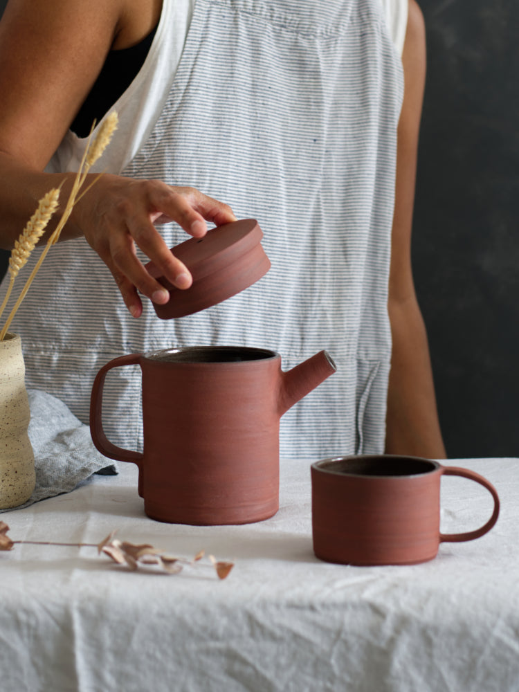 Hand die deksel van rode theepot vasthoudt. Op tafel een rode mok van dezelfde klei. 