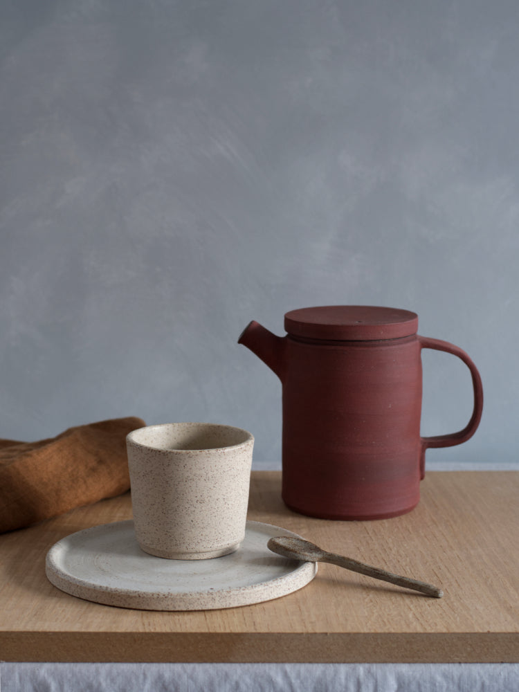 Handgemaakte rode theepot van steengoed houten plank naast licht gespikkeld kopje en een cakebord lichtgespikkeld met keramiek lepeltje