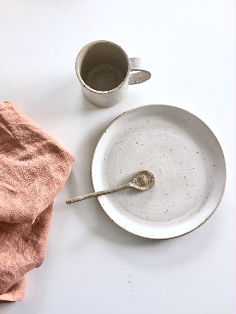 Handgemaakt dinerbord van keramiek met mok, keramieken lepel en theedoek ernaast op een witte achtergrond
