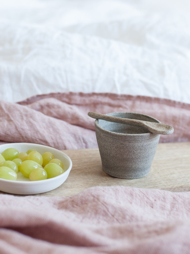 handgemaakt bruin kopje op een houten dienblad met druiven ernaast op een wit en roze fluffy dekbed