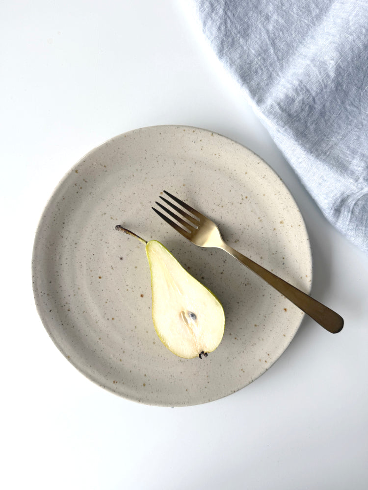Beige dinerbord handgemaakt met een halve peer erop en een goudkleurige vork, blauwe theedoek bovenaanzicht