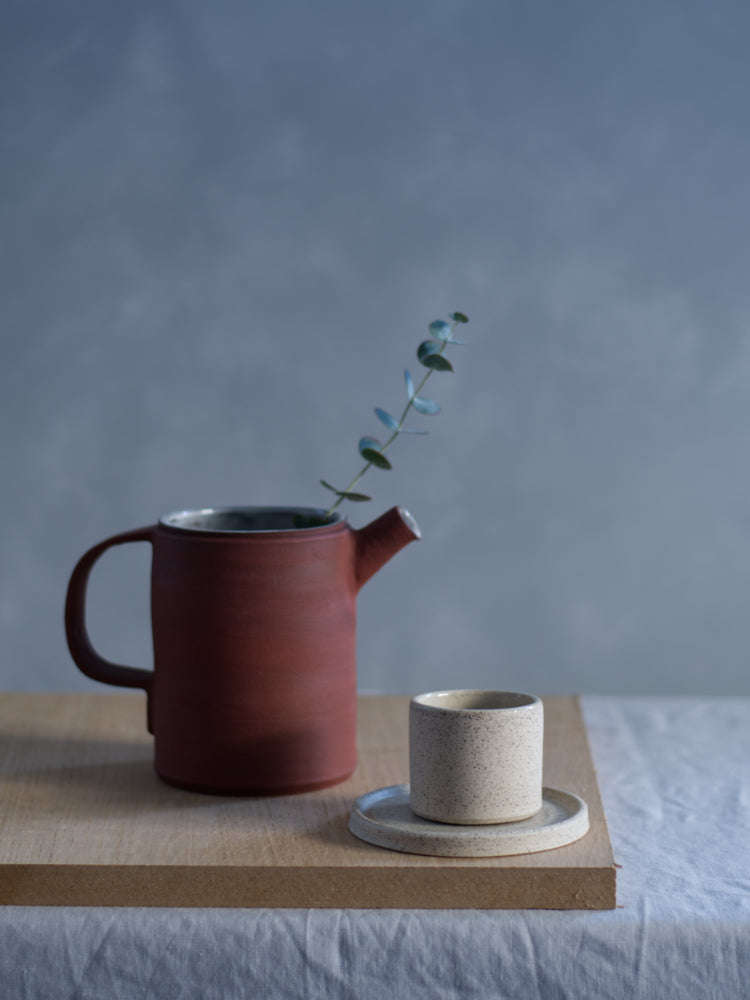 Handgemaakt espressokopje van gespikkelde klei op een handgemaakt schoteltje naast een theepot van rood steengoed grijze achtergrond
