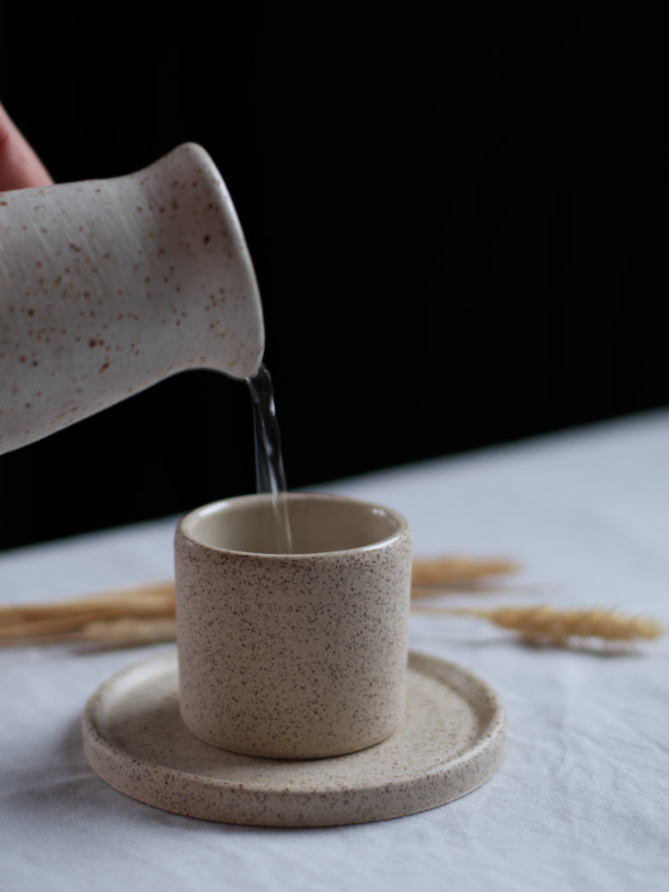 Handgemaakt espressokopje op een bijpassend schoteltje  waarin water wordt geschonken uit een whitewashed schenkkannetje op een lichtgrijs tafelkleed