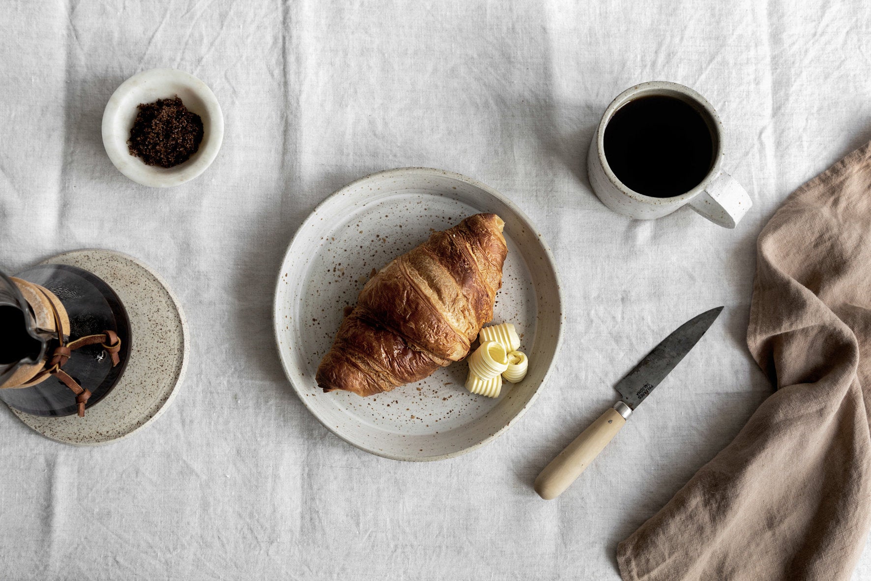 Gedekte tafel met handgemaakt servies, croissant met boter en koffiepot, bovenaanzicht.