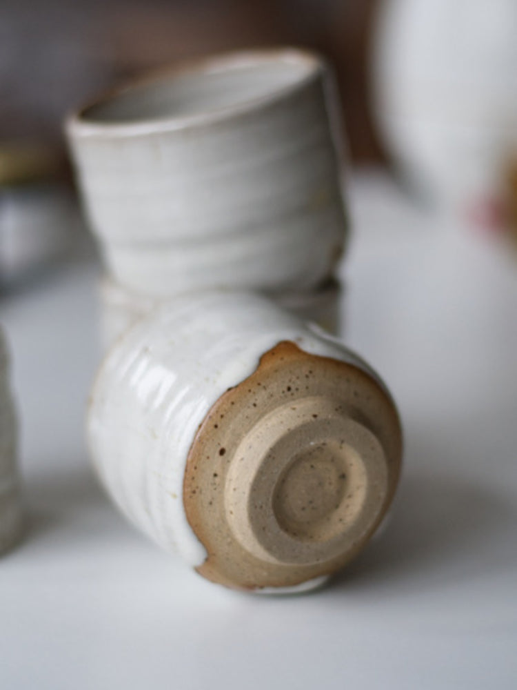 Handgemaakt keramieken kopje met wit glazuur en spikkels liggend zodat de onderkant zichtbaar is, met andere kopjes vaag erachter