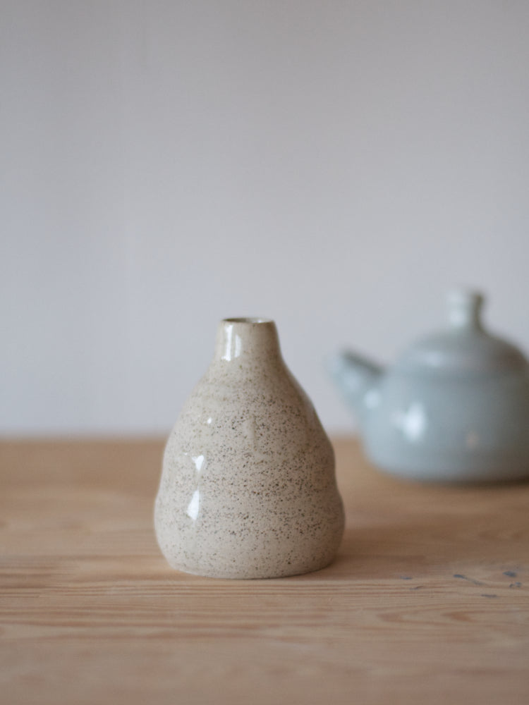 Handgemaakt klein vaasje, gemaakt van gespikkelde klei met licht golvende vorm naast een klein blauw theepotje