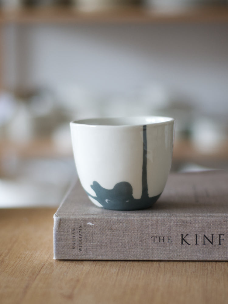 Handgemaakte Waterlijn kopje Large van wit porselein met een donkergrijze  waterlijn, staand op een boek met linnen kaft, op een houten tafel
