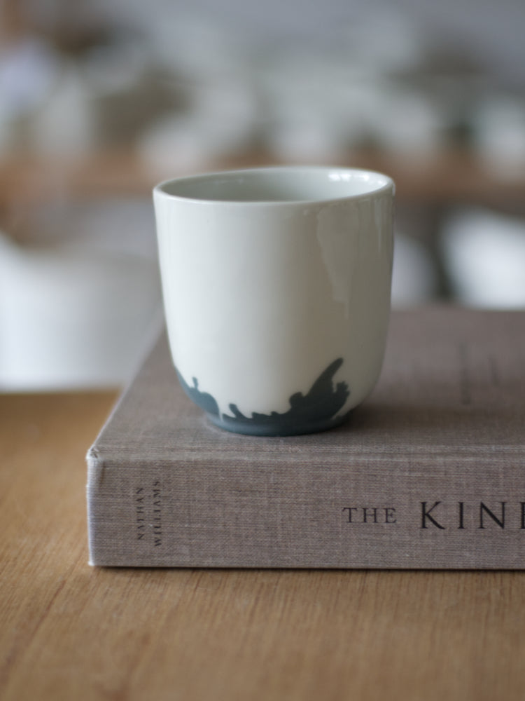 Handgemaakte Waterlijn kopje large van wit porselein met een donkergrijze waterlijn, staand op een boek met linnen kaft, op een houten tafel