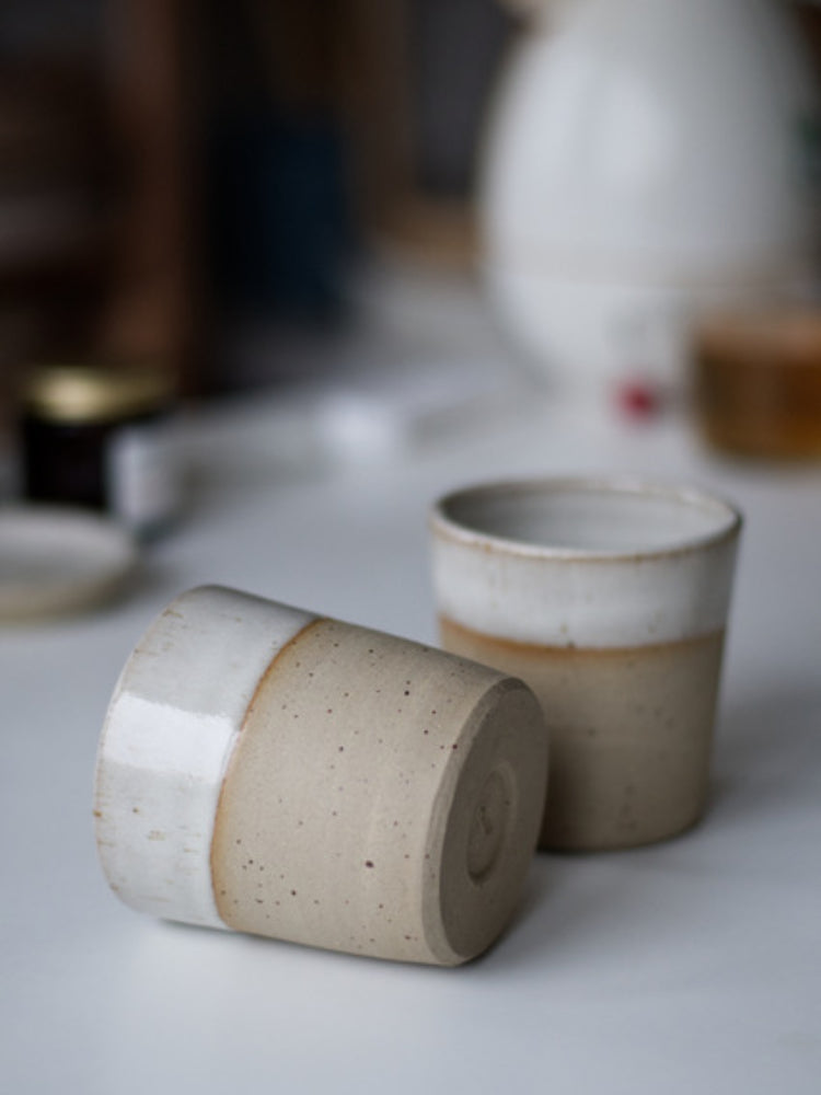 Handgemaakte beker van gespikkeld steengoed met glanzend wit glazuur waarbij de spikkels van de klei zichtbaar blijven en alleen de bovenrand geglazuurd is. Een beker ligt op zijn zij.