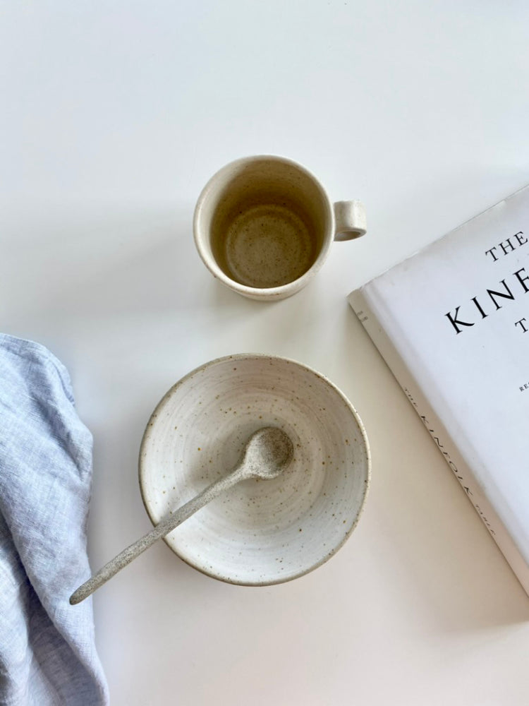 Flatlay van een handgemaakte whitewashed kom met keramieken lepel, bijpassende mok, theedoek en boek op houten ondergrond – handgemaakt keramiek voor slow living.