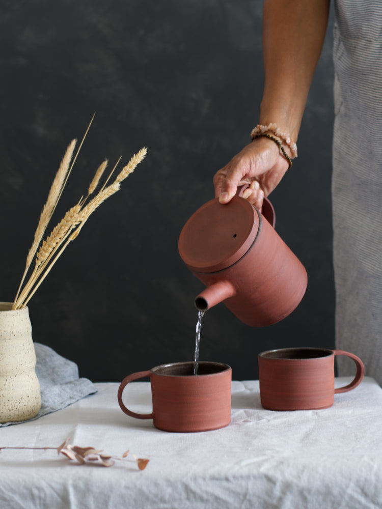 Handgemaakte rode keramieken mokken met een hand die met een rode theepot schenkt in de mokken