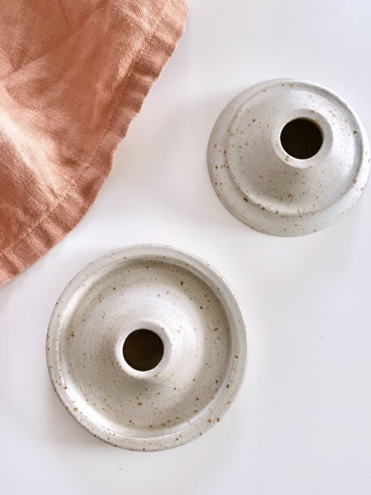 Handgemaakte whitewashed kandelaars van keramiek op een witte achtergrond met een theedoek ernaast.