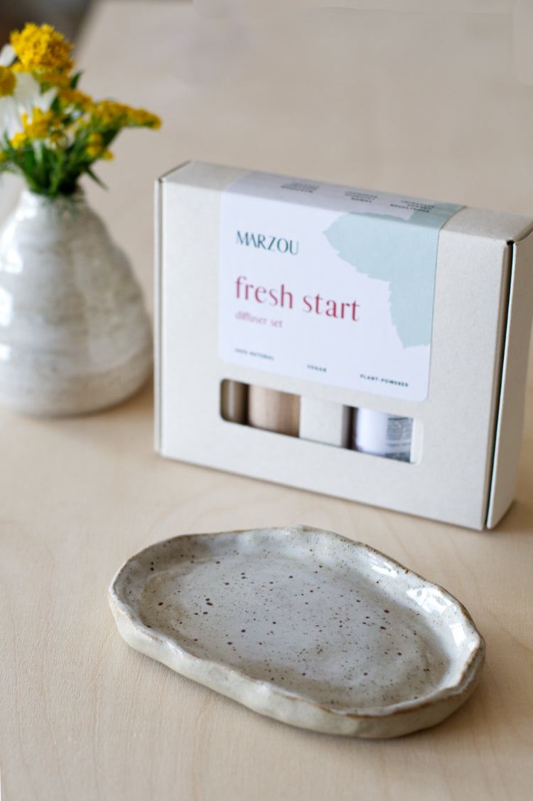 Handgemaakt keramisch schoteltje met geurolie en houten diffuser naast een vaasje met gele bloemen