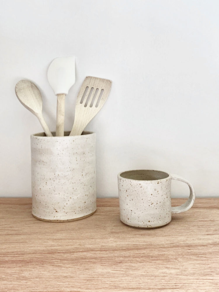 Keukengerei houder van keramiek met spatels en een mok ernaast op een houten tafelblad.