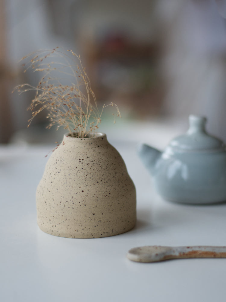 Handgemaakt klein vaasje van gespikkelde klei ongeglazuurde buitenkant en in de achtergrond een klein blauw theepotje op witte tafel
