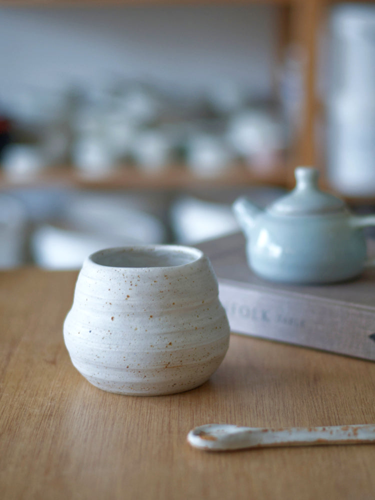 Handgemaakt klein vaasje van gespikkelde klei mat wit glazuur en in de achtergrond een boek met een klein blauw theepotje erop