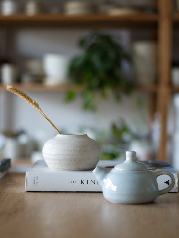 Handgemaakt vaasje met wit mat glazuur, op een boek, en klein blauw theepotje ervoor