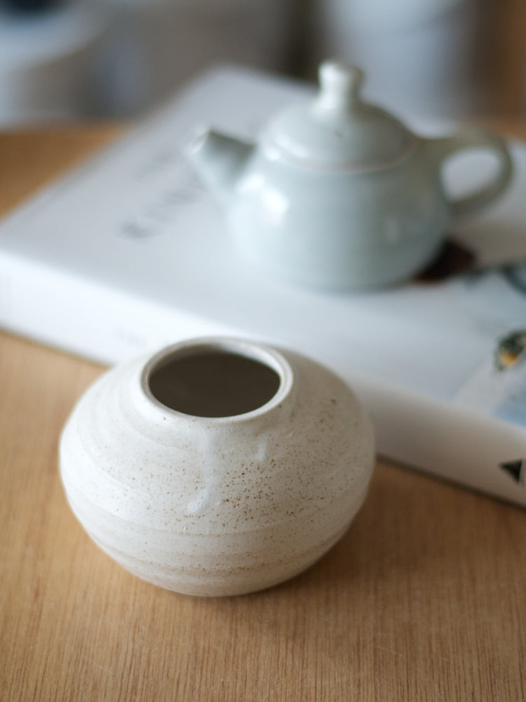Handgemaakt vaasje met wit mat glazuur, met vaag erachter, op een boek, een klein blauw theepotje