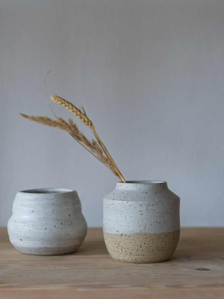 Handgemaakt vaasje van gespikkelde klei half wit mat geglazuurd naast een ander mat wit geglazuurd vaasje. 