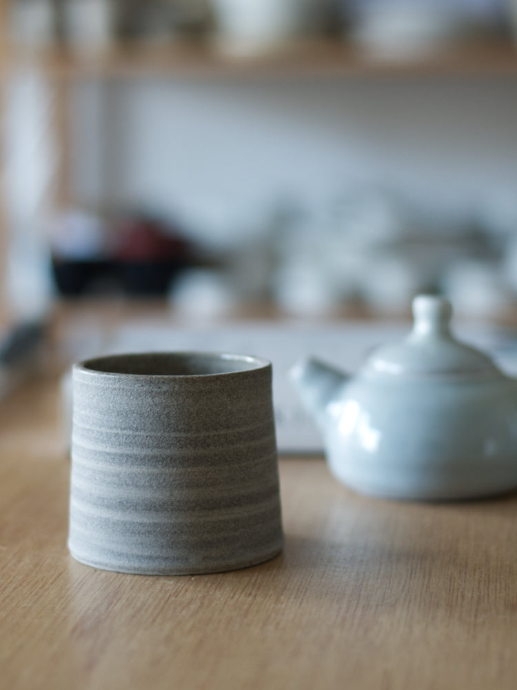 Handgemaakt kopje van donkere klei met ongeglazuurde buitenkant, met vaag een blauw theepotje op de achtergrond