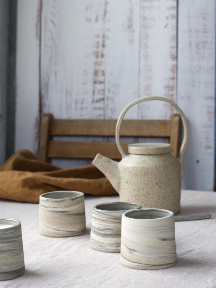 Verschillende handgemaakte landschapskopjes, gedraaid van 3 soorten klei op een tafel met tafelkleed en een theepot, slow living vibe