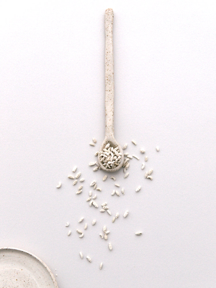 Lange beige gespikkelde handgemaakte lepel van keramiek met rijstkorrels en een bordje, stilleven