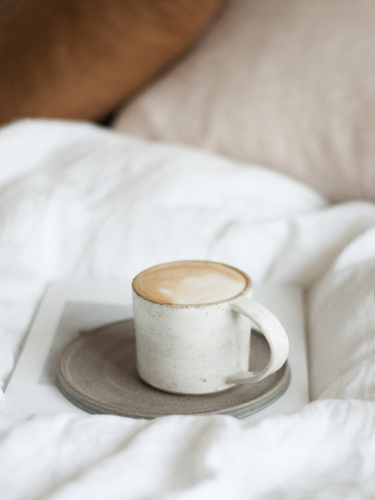 whitewashed mok met glanzend glazuur op een handgemaakt donker keramieken cakebord op een fluffy wit linnen dekbed 
