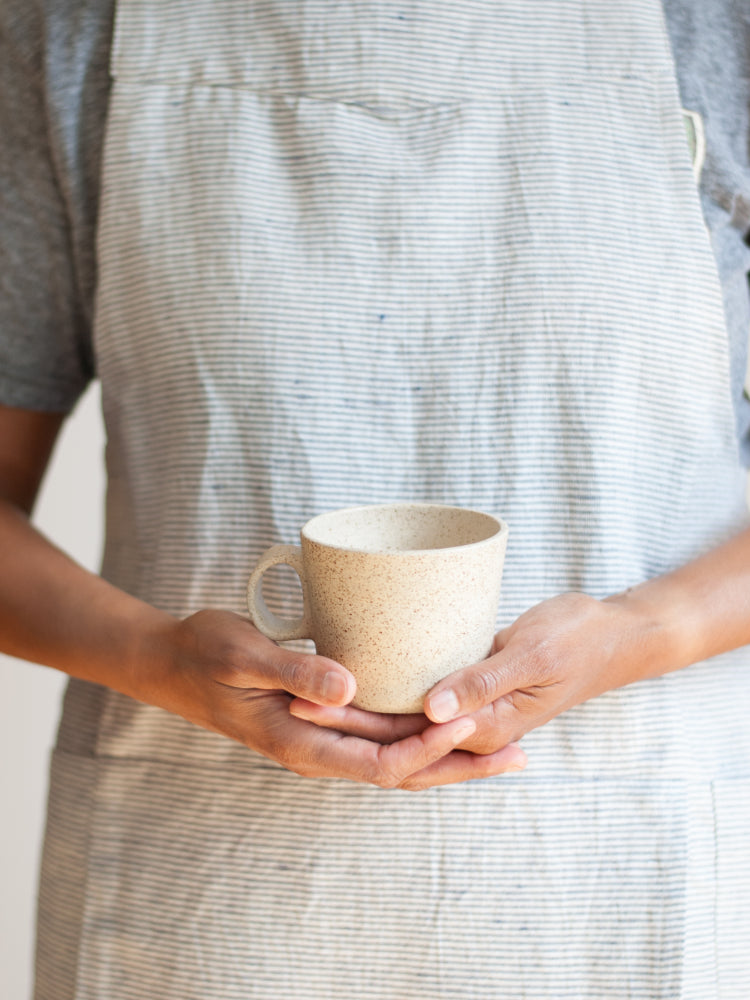 Twee handen voor een schort die een lichtgespikkelde handgemaakte mok vasthouden