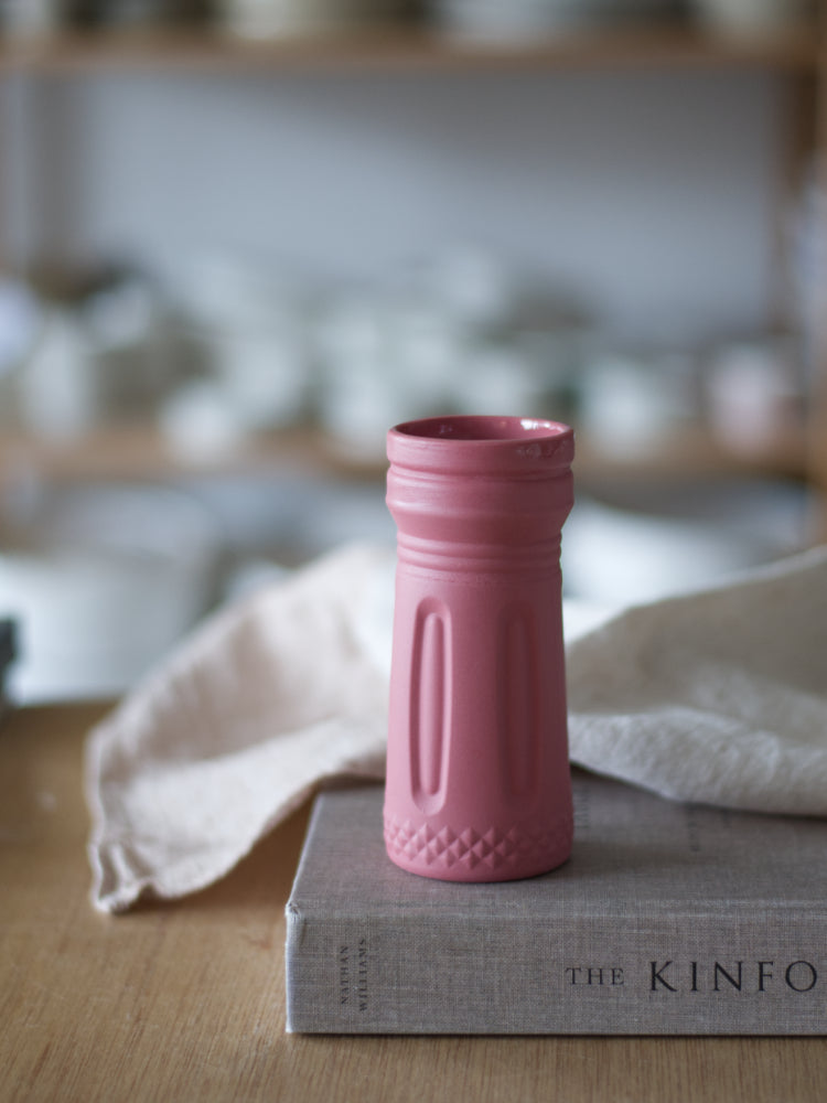 Handgemaakt rood porseleinen vaasje met boogpatroon en ruitjes op houten tafel naast een boek en theedoek