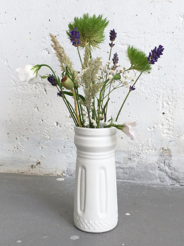 Handgemaakt wit porseleinen vaasje met bloemen en grassen voor witte muur en op industriële grijze betonnen vloer