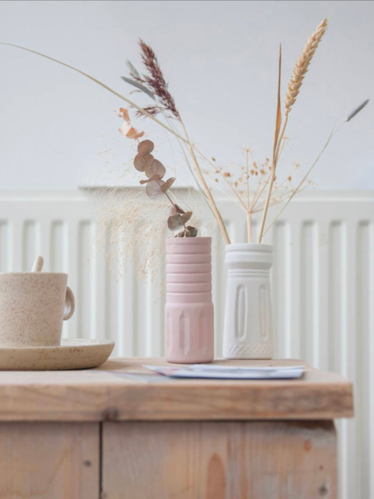 Twee handgemaakte porseleinen vaasjes met grassen en droogbloemen erin. Op een houten kruk